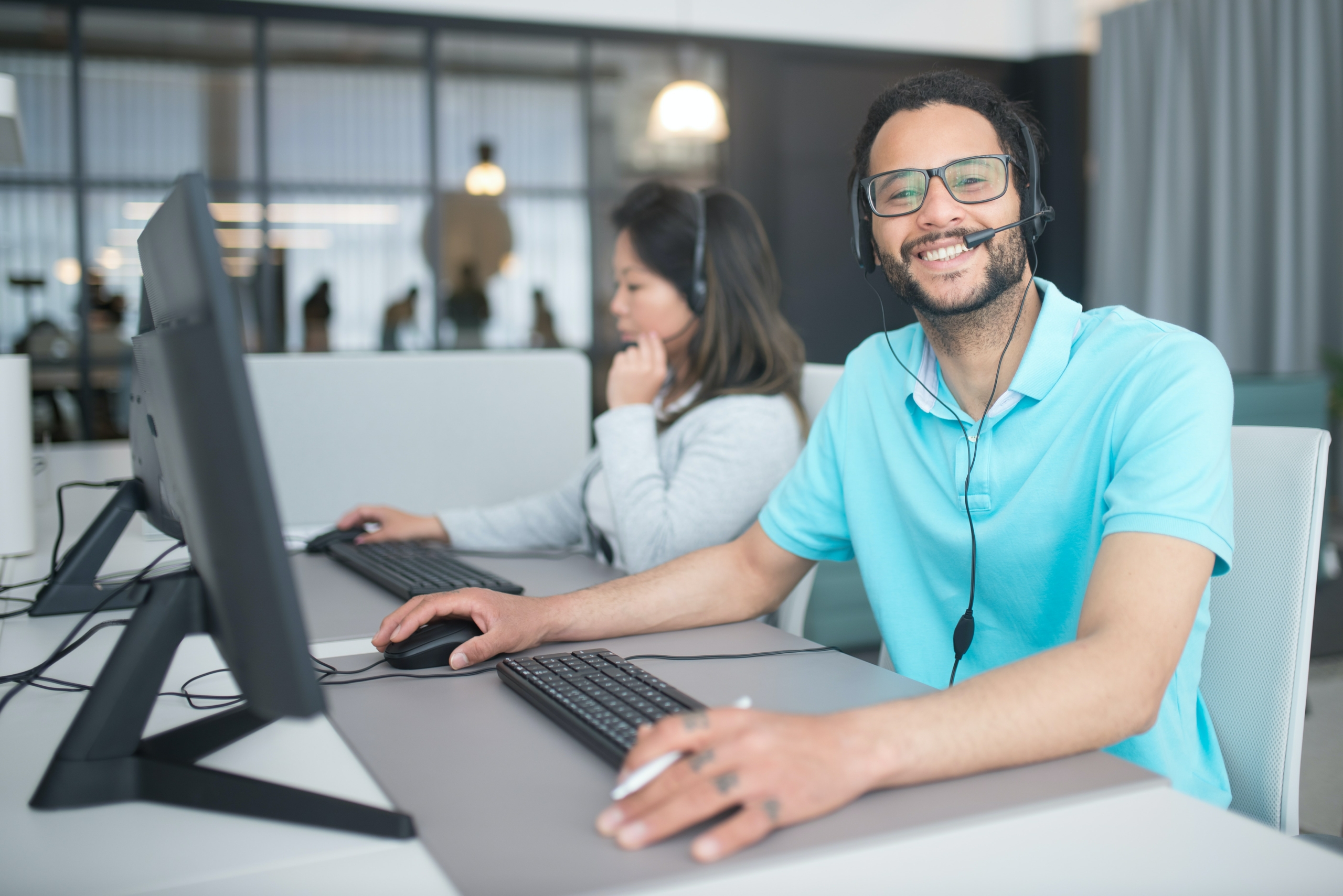 A photo of a man wearing a phone headset and sitting at a computer, smiling at the viewer of the photo.